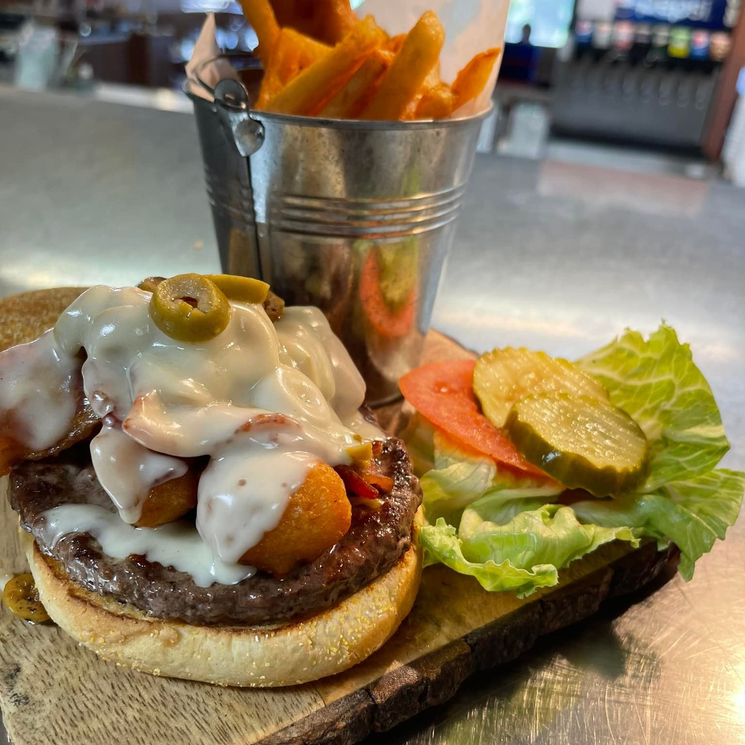 A burger with various topping but no top bun, with other toppings placed next to it on a piece of wood. Behind it is a small metal pail with fries in it.