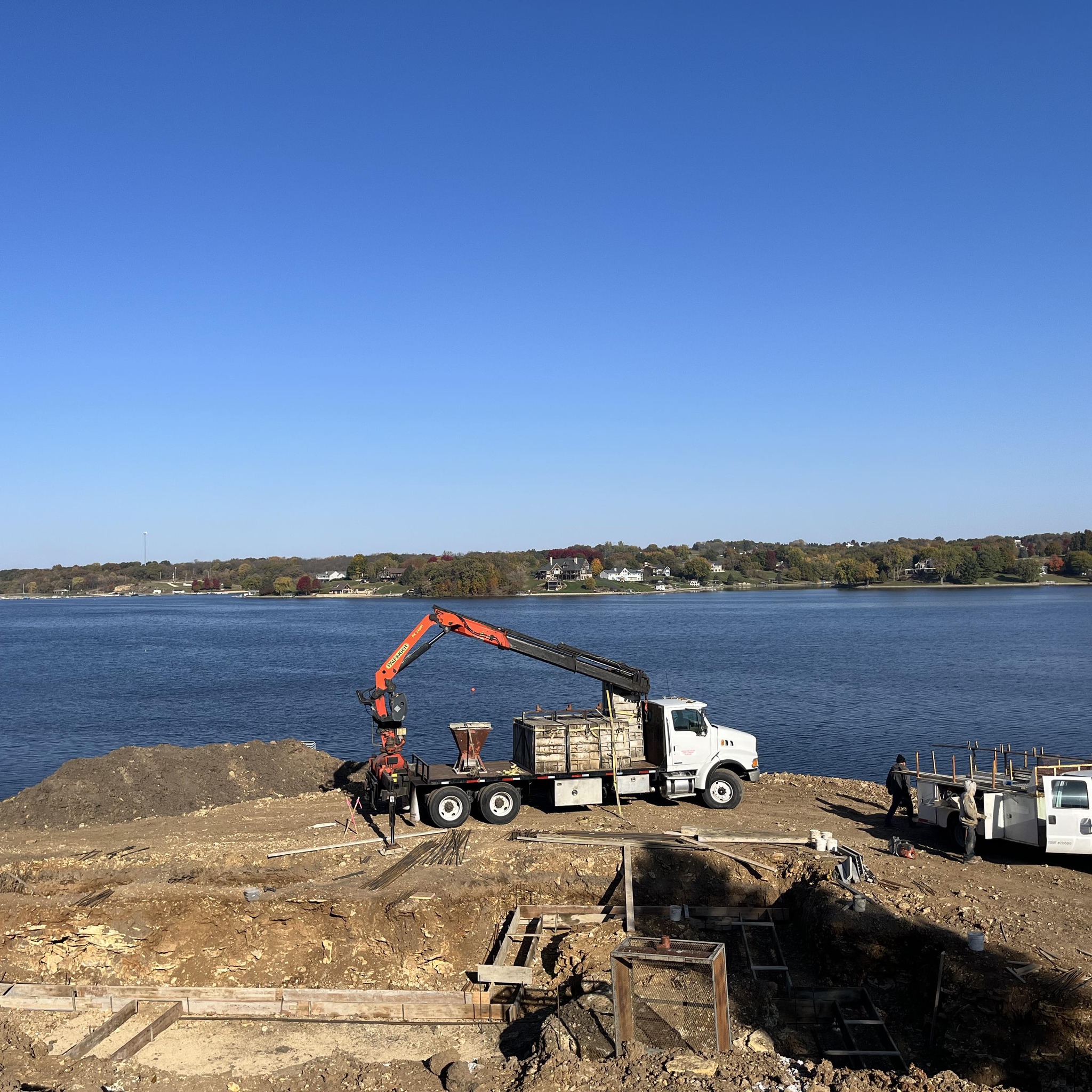 Construction site with vehicles near a body of water with houses seen on the other side of the water in the distance.