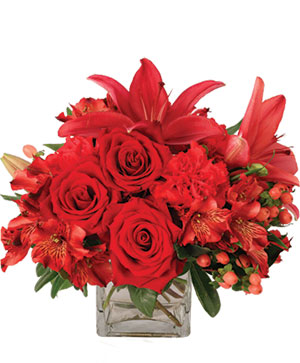 A small glass vase with roses, lilies and other red flowers in it.