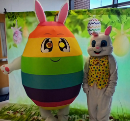 Two people in easter costumes, one a rabbit and the other a rainbow egg in front of an easter themed backdrop.